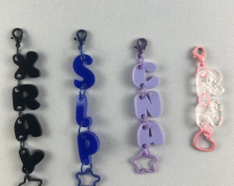 Puede incluir: Cuatro llaveros coloridos con diferentes nombres escritos en letras de burbuja. Los llaveros son negro, azul, morado y transparente con acentos rosados. Cada llavero tiene un encanto de estrella en la parte inferior.