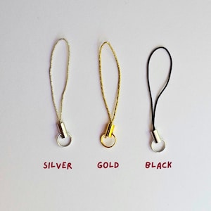 May include: Three phone charms in silver, gold, and black. Each charm has a loop and a metal cylinder. The words "SILVER", "GOLD", and "BLACK" are written below each charm.