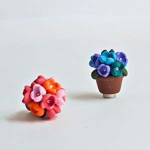 May include: Two miniature floral sculptures. One is a bouquet of pink, orange, and red flowers. The other is a potted plant with blue, purple, and green flowers. Both have small, sparkly accents. The background is white.