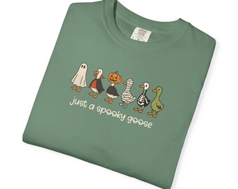 May include: Sage green t-shirt featuring a line of cartoon geese dressed in Halloween costumes, including a ghost, vampire, pumpkin, mummy, skeleton, and a green goose. The text "just a spooky goose" is printed below the geese.