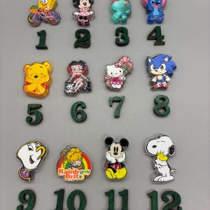 May include: A collection of colorful character-themed charms, including SpongeBob, Minnie Mouse, Stitch, Winnie the Pooh, Betty Boop, Hello Kitty, Sonic, Lumiere, Rainbow Brite, Mickey Mouse, and Snoopy. Each charm is paired with a corresponding green wooden number.