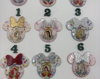 May include: Nine Minnie Mouse-shaped resin embellishments with princess portraits. Each has a glittery bow and a different princess image. The embellishments are numbered 1 through 9 with green numbers.