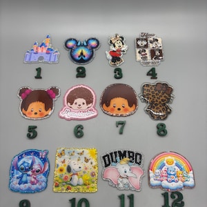 May include: A collection of twelve colorful character-themed embellishments. Designs include a castle, Mickey Mouse, Minnie Mouse, Dumbo, Stitch, Snoopy, and other cartoon characters. Each embellishment has a glittery border.