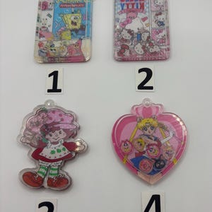 May include: Four clear plastic keychains with cartoon characters. The keychains feature SpongeBob SquarePants, Hello Kitty, Strawberry Shortcake, and Sailor Moon. Each keychain has a small loop at the top for attaching to keys or a bag.