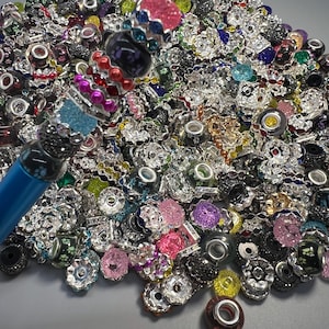 May include: A collection of colorful beads, including silver, black, pink, and blue, with various shapes and designs. Some beads are embellished with rhinestones. A blue pen is visible in the image, with a decorative top featuring pink and blue accents.