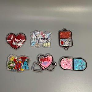 May include: A collection of six medical-themed badge reels. The reels feature various designs, including a heart with an EKG line, a pharmacy tech design, a blood bag, a CMA design, a stethoscope heart, and a pill.