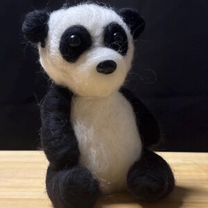 May include: A handmade panda bear figurine with black and white wool. The panda has black ears, eyes, and limbs, with a white face and body. The panda is sitting on a wooden surface against a black background.