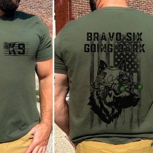 May include: A green t-shirt with a black and white American flag design featuring a German Shepherd wearing a helmet and night vision goggles. The text "BRAVO SIX GOING BARK" is printed on the back of the shirt. The front of the shirt has a black and white American flag design with the text "K9" printed on it.