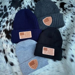May include: Four knitted beanies in different colors, each with a leather patch featuring an American flag design. The beanies are gray, navy blue, black, and light gray.