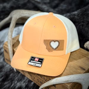 May include: A peach-colored trucker hat with a white mesh back. The hat features a leather patch of the Montana state outline with a heart design. The Richardson brand label is visible on the front.