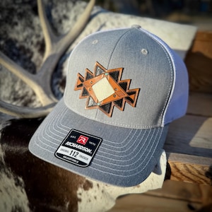 May include: A gray trucker hat with a white mesh back and a decorative patch. The patch features a geometric design in brown, black, and tan, with small turquoise accents. The hat has a curved brim and an adjustable strap.