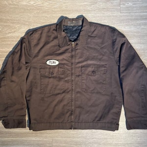 May include: A dark brown work jacket with a zipper closure and a white patch that says "Mike" on the left chest.