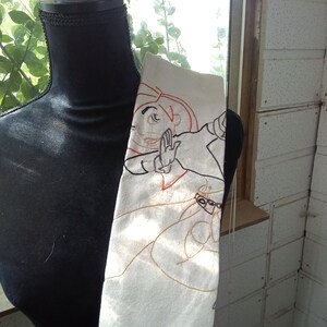 Op de afbeelding: Een witte rechthoekige theedoek met een gedetailleerde zwarte en oranje geborduurde omtrek van het animatiepersonage Kim Possible in een dynamische actiehouding, weergegeven op een zwarte etalagepop-torso voor een helder raam.