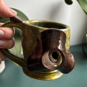 May include: A handmade ceramic mug with a unique design. The mug is primarily green and brown with a sculpted brown eye on the side. The handle is also brown. The mug is held by a person.