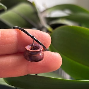 May include: A miniature ceramic pot pendant with a dark purple glaze and a brown rim, suspended from a black cord. The small pot is held against a backdrop of green leaves, showcasing its handcrafted details.