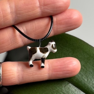 May include: A small, ceramic goat pendant with black and brown markings, suspended from a black cord necklace. The goat figurine is white with brown spots and black hooves. The necklace is displayed against a blurred background.