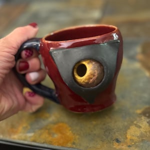 May include: A glossy red ceramic mug featuring a dark blue handle and a prominent, three-dimensional sculpted eye element. The eye is set within a black triangular frame, showcasing a yellow iris and a dark pupil, creating a distinct focal point.