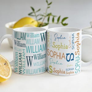 Può includere: Due tazze in ceramica bianca con nomi personalizzati. Una tazza presenta il nome "William" in varie tonalità di blu, mentre l'altra mostra "Sophia" in un mix di colori. Sono visibili anche un limone tagliato a metà e una teiera.