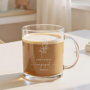 Puede incluir: Una taza de vidrio transparente llena de café y cubierta con una capa de espuma. La taza tiene un asa y presenta las palabras "JIMMY & NYAH engaged 10.10.25" junto con un diseño botánico. La taza está sobre una mesa blanca.