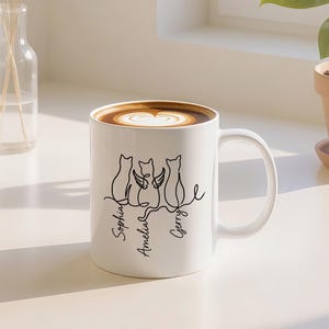 以下が含まれることがあります： 3匹の猫の黒い線画とSophia、Amelia、Gerryの名前が描かれた白いセラミックマグ。マグカップにはコーヒーが入り、ハート型のデザインが施されています。マグカップは白い表面に置かれています。