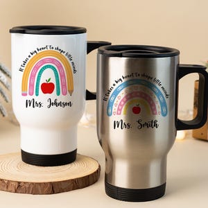 Puede incluir: Dos tazas de viaje, una blanca y otra plateada, con tapas y asas negras. Cada taza presenta un dise&ntilde;o de arco&iacute;ris con una manzana y un l&aacute;piz, y el texto "It takes a big heart to shape little minds" y un nombre.