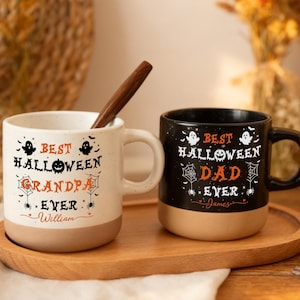 May include: Two ceramic mugs with a two-tone design. The left mug is cream with the text "BEST HALLOWEEN GRANDPA EVER" and "William". The right mug is black with the text "BEST HALLOWEEN DAD EVER" and "James". Both feature ghost and spiderweb graphics.