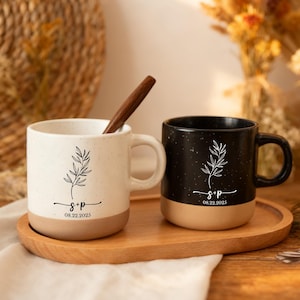 Puede incluir: Dos tazas de cerámica, una crema y otra negra, con acabado moteado y base marrón, sobre una bandeja de madera. Cada taza presenta un diseño botánico negro, iniciales y la fecha "08.22.2023". Una cuchara de madera está en la taza crema.