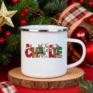 Könnte beinhalten: Eine weiße Emaille-Tasse mit silbernem Rand, die den Namen "Charlie" in einem festlichen, weihnachtlichen Design zeigt. Die Buchstaben sind mit Weihnachtsmützen, Rentiergeweihen und anderen Feiertagselementen verziert. Die Tasse steht auf einem Holzuntersetzer.