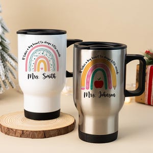 Puede incluir: Dos tazas de viaje con tapas y asas negras. Una es blanca, la otra de acero inoxidable. Ambas presentan un dise&ntilde;o de arco&iacute;ris, el texto "It takes a big heart to shape little minds" y un nombre. La taza blanca est&aacute; sobre un posavasos de madera.
