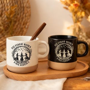 Può includere: Due tazze in ceramica, una bianca maculata e una nera, entrambe con base color cuoio e una grafica nera di tre streghe che danzano in cerchio sotto la scritta God Forbid Women Have Hobbies. Un cucchiaio di legno è inserito nella tazza bianca.