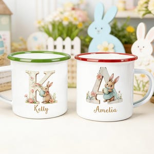 Puede incluir: Dos tazas de esmalte blanco con diseños de Pascua. Una taza tiene un borde verde y presenta la letra "K" con un conejito y el nombre "Kelly". La otra tiene un borde rojo, la letra "A", un conejito y el nombre "Amelia".