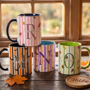May include: Four ceramic mugs with vertical stripes and large letters. One mug is pink with the letter R, one is orange and black with M, one is purple and orange with N, and one is green and white with O. A round wooden coaster reads Hilda.