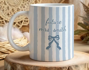 Könnte beinhalten: Weiße Keramiktasse mit blauen vertikalen Streifen und dem Text "future mrs. smith" in Schreibschrift. Unter dem Text befindet sich eine blaue Schleifengrafik. Die Tasse steht auf einer Holzscheibe.