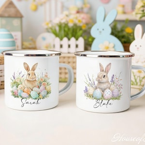 Könnte beinhalten: Zwei weiße Emaille-Tassen mit silbernen Rändern und Griffen. Jede Tasse zeigt eine Aquarell-Illustration eines Hasen, umgeben von Ostereiern und Lavendel. Eine Tasse ist mit "Sarah" beschriftet, die andere mit "Blake". Osterdekoration im Hintergrund.