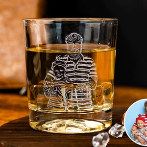 Op de afbeelding: Een helder whiskyglas gevuld met amberkleurige vloeistof, met een geëtste afbeelding van een persoon en een kind. Het glas staat op een donkere houten onderzetter, met ijsblokjes. Een kleine, ronde foto staat op de achtergrond.