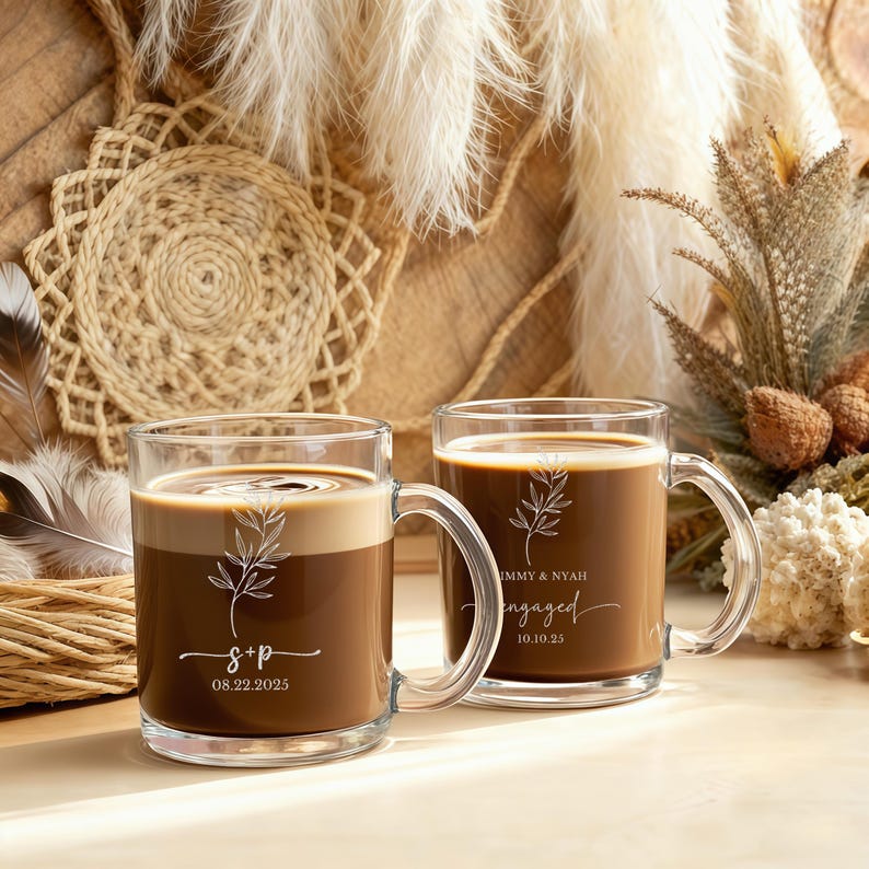 Peut inclure: Deux mugs en verre transparent remplis de caf&eacute;, avec un motif de feuille d&eacute;coratif et un texte personnalis&eacute;. L'un des mugs porte l'inscription "S&P" avec une date, l'autre "Jimmy & Nyah engaged 10.10.23". Les mugs ont une anse et sont pos&eacute;s sur un fond neutre.