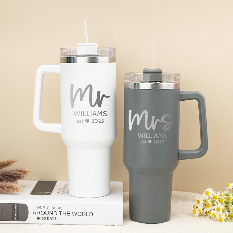 Pu&ograve; includere: Due tazze da viaggio in acciaio inox personalizzate con coperchi e cannucce. La tazza bianca dice "Mr. Williams est. 2025" e la tazza grigia dice "Mrs. Williams est. 2025".