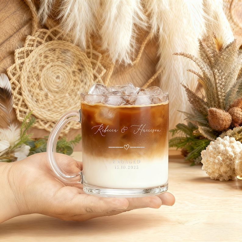 Peut inclure: Une tasse en verre transparente remplie de caf&eacute; glac&eacute;, tenue dans une main. La tasse a une anse et porte les noms "Rebecca & Harrison" en &eacute;criture cursive, une ligne avec un c&oelig;ur et le mot "ENGAG&Eacute;" avec la date 15.10.2025.
