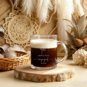 Peut inclure: Un mug en verre transparent rempli de caf&eacute; noir et surmont&eacute; d'une couche de cr&egrave;me. Le mug a une anse et porte les inscriptions "JIMMY & NYAH" et "engaged 10.10.25" en &eacute;criture &eacute;l&eacute;gante. Le mug est pos&eacute; sur un dessous de verre en bois.