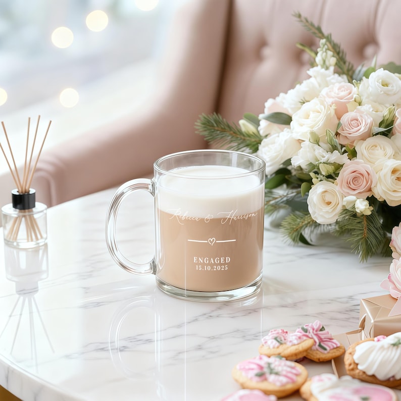 Peut inclure: Mug en verre transparent rempli de caf&eacute; au lait, avec l'inscription "Rebecca & Harrison", un c&oelig;ur et "ENGAG&Eacute;S 15.10.2025". Le mug est pos&eacute; sur une surface en marbre, &agrave; c&ocirc;t&eacute; d'un bouquet de roses blanches et roses.