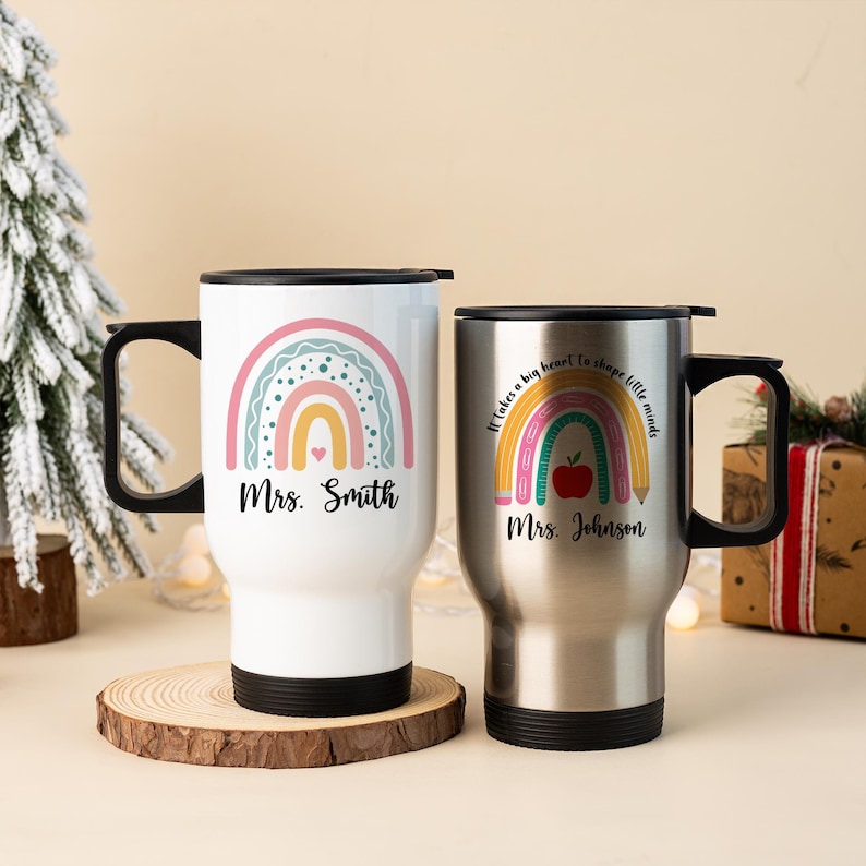 Puede incluir: Dos tazas de viaje con asas y tapas negras. Una es blanca con un dise&ntilde;o de arco&iacute;ris y el nombre "Mrs. Smith". La otra es plateada con un dise&ntilde;o de arco&iacute;ris, manzana y l&aacute;piz, y el nombre "Mrs. Johnson". Ambas tienen bases negras.