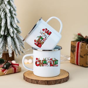 K&ouml;nnte beinhalten: Zwei wei&szlig;e Emaille-Tassen mit silbernen R&auml;ndern. Die obere Tasse zeigt das Wort BELIEVE in roten, gr&uuml;nen und wei&szlig;en Buchstaben im Feiertagsdesign. Die untere Tasse zeigt den Namen CHARLIE in &auml;hnlicher, festlicher Schriftart.