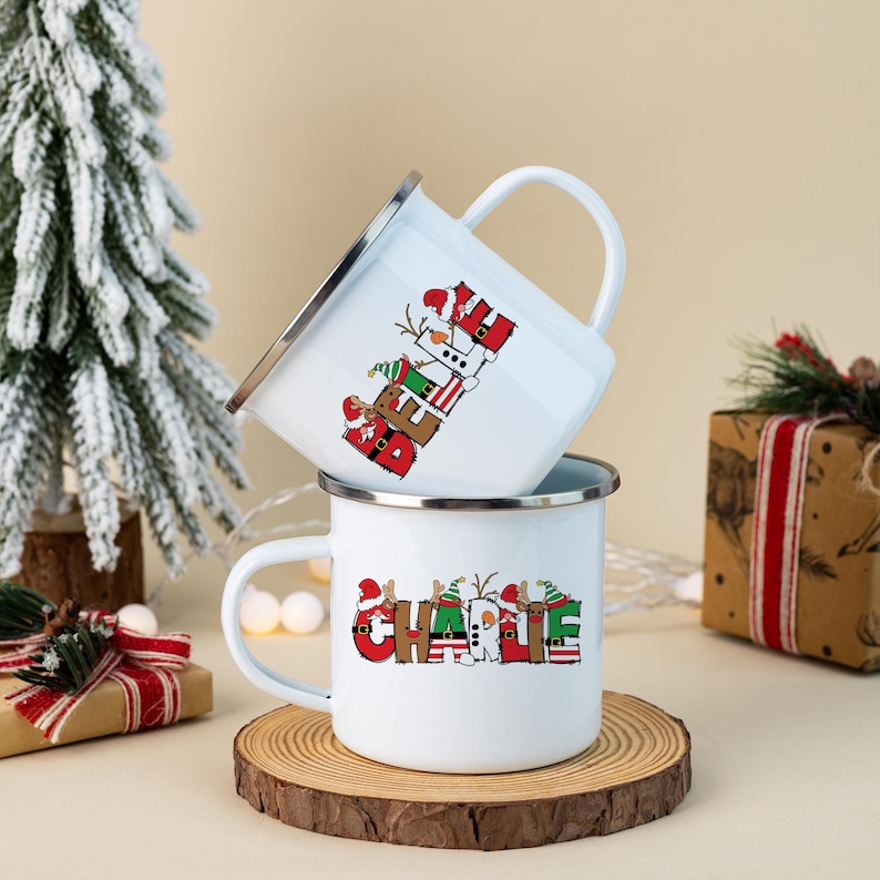 K&ouml;nnte beinhalten: Zwei wei&szlig;e Emaille-Tassen mit silbernen R&auml;ndern. Die obere Tasse zeigt das Wort BELIEVE in roten, gr&uuml;nen und wei&szlig;en Buchstaben im Feiertagsdesign. Die untere Tasse zeigt den Namen CHARLIE in &auml;hnlicher, festlicher Schriftart.