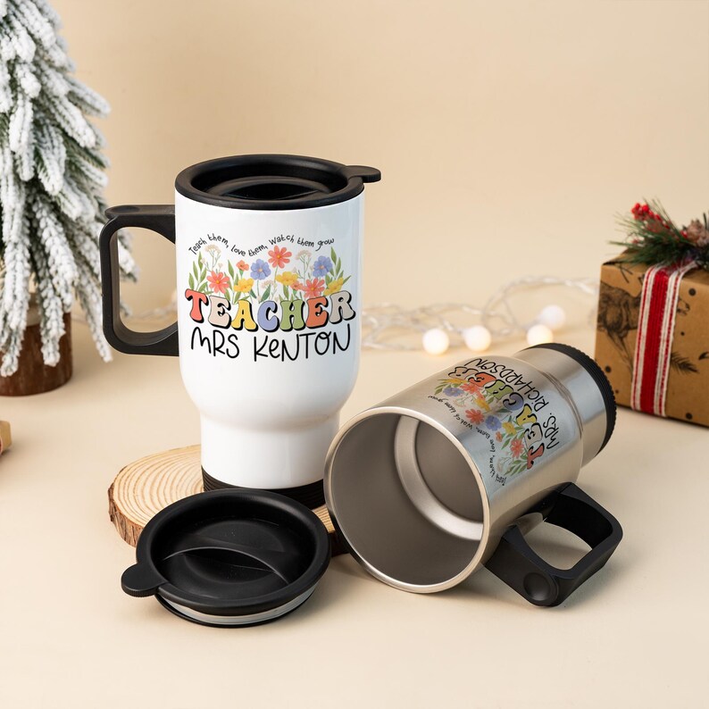 May include: Two travel mugs, one white and one silver, with black handles and lids. The white mug has the text "TEACHER MRS KENTON" with floral accents. The silver mug has the text "MRS. RICHARDSON" with floral accents. The mugs are on a wooden coaster.