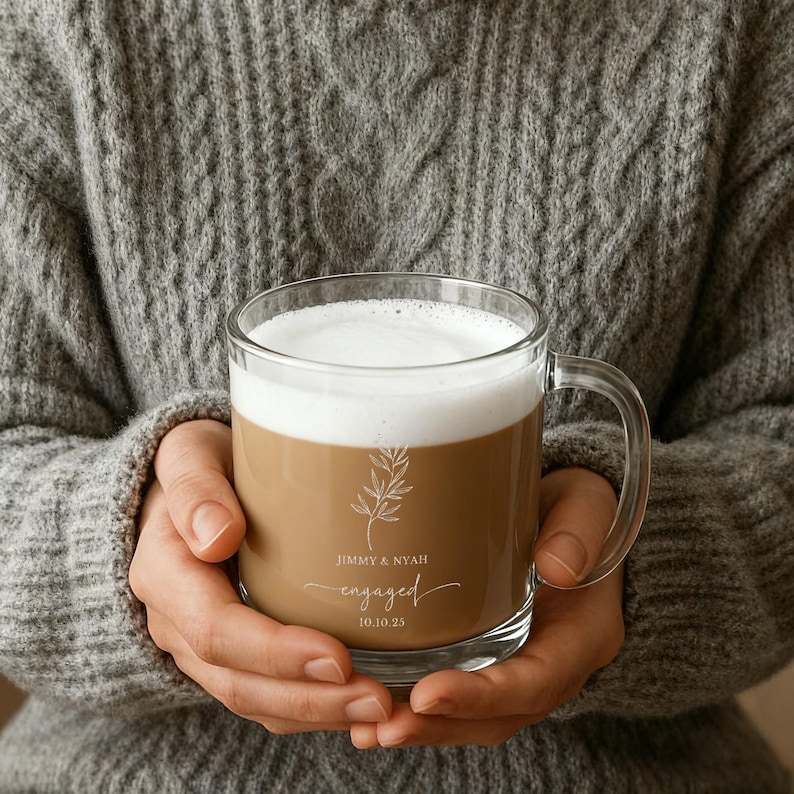 Peut inclure: Une tasse en verre transparent remplie d'une boisson au caf&eacute; et recouverte de mousse blanche. La tasse a une anse et porte les mots "Jimmy & Nyah engaged 10.10.25" ainsi qu'un motif botanique. La tasse est tenue par une personne portant un pull gris.