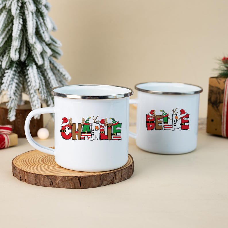 K&ouml;nnte beinhalten: Zwei wei&szlig;e Emaille-Tassen mit silbernen R&auml;ndern. Die vordere Tasse zeigt den Namen CHARLIE in roten, gr&uuml;nen und wei&szlig;en weihnachtlichen Blockbuchstaben mit Weihnachtsmannm&uuml;tzen und Rentiergeweihen. Die hintere Tasse zeigt den Namen BELLE.