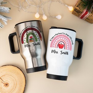 Puede incluir: Dos tazas de viaje, una plateada y otra blanca, con asas negras. Cada taza presenta un dise&ntilde;o de arco&iacute;ris con una manzana y el texto "Se necesita un gran coraz&oacute;n para dar forma a las mentes peque&ntilde;as" y "Sra. Smith."