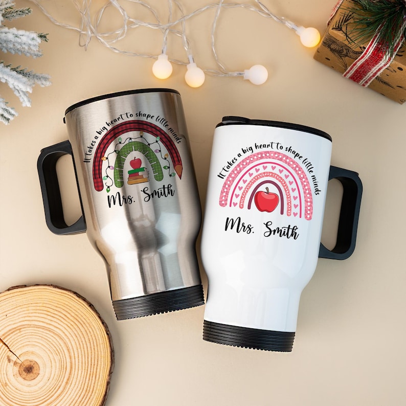 Puede incluir: Dos tazas de viaje, una plateada y otra blanca, con asas negras. Cada taza presenta un dise&ntilde;o de arco&iacute;ris con una manzana y el texto "Se necesita un gran coraz&oacute;n para dar forma a las mentes peque&ntilde;as" y "Sra. Smith."