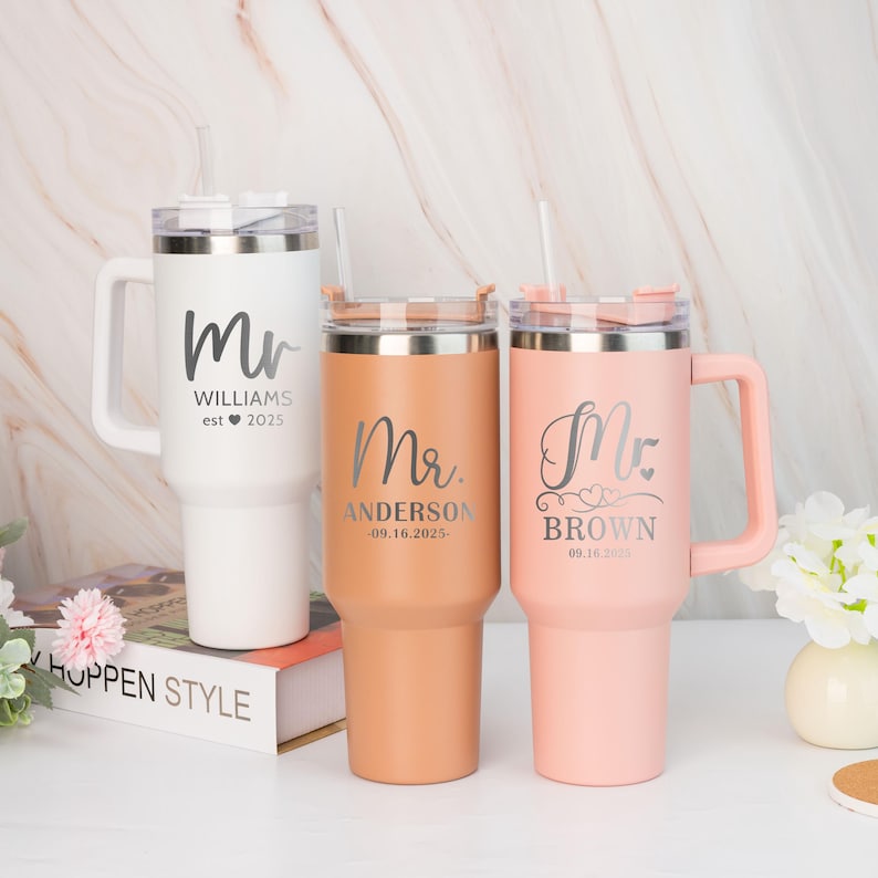 Pu&ograve; includere: Tre bicchieri in acciaio inossidabile personalizzati con coperchi e manici. I bicchieri sono bianco, marrone e rosa. I bicchieri sono incisi con il testo "Mr. Williams est. 2025", "Mr. Anderson 09.16.2025" e "Mr. Brown 09.18.2025".