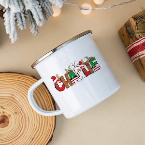K&ouml;nnte beinhalten: Ein wei&szlig;er Emaillebecher mit silbernem Rand, der den Namen CHARLIE in gro&szlig;en, bunten Buchstaben zeigt. Jeder Buchstabe ist mit weihnachtlichen Motiven verziert, darunter Rentiere, Weihnachtsm&uuml;tzen, Streifenmuster und ein Schneemann.
